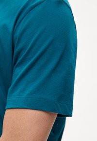 Άνω μπράτσο ατόμου που φοράει ένα τιρκουάζ βαμβακερό κοντομάνικο μπλουζάκι, με φόντο έναν απλό ανοιχτόχρωμο τοίχο.