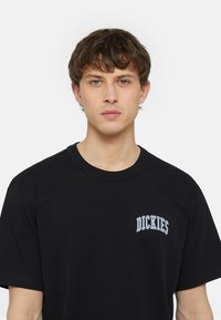 Fekete, rövid ujjú pamut póló, fehér "DICKIES" logóval hímzéssel a mellrészen, kerek nyakkivágással és laza szabással.