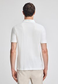 Camisa polo blanca de manga corta con cuello clásico, confeccionada en una tela suave. Presenta un dobladillo recto y sutiles detalles de costura en las mangas.