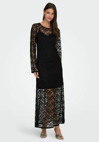 Czarna koronkowa sukienka z długimi rękawami, przezroczyst�ą warstwą wierzchnią, dopasowaną podszewką, kwiatowym wzorem i rozkloszowanym dołem. Do kompletu sandały na cienkich, metalicznych paseczkach.