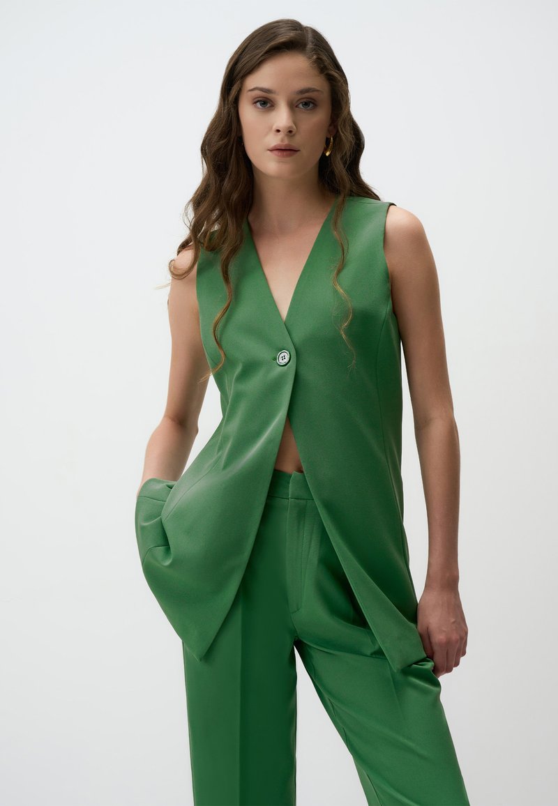 Blazer verde senza maniche con un profondo scollo a V e un dettaglio con bottone, abbinato a pantaloni a gamba larga coordinati in un tessuto liscio e opaco.