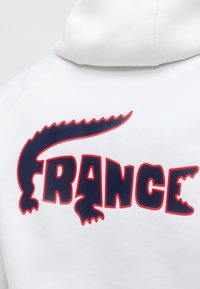Λευκή φούτερ με κουκούλα με στιλιζαρισμένο σκοτεινό μπλε και κόκκινο κείμενο "FRANCE", που περιλαμβάνει σχήμα κροκοδείλου γύρω από τα γράμματα στην πλάτη.