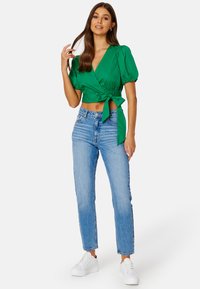 Top avvolgente verde corto con maniche a sbuffo, legato in vita, abbinato a jeans azzurri a vita alta e sneaker bianche con plateau.