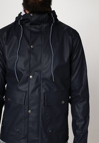 Marinblå vattentät jacka med frontdragkedja, dragsko i huvan, två stora framfickor och silverfärgade tryckknappar. Slät, glänsande yta.