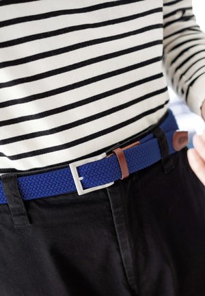 Persona che indossa pantaloni neri, una maglia a maniche lunghe bianca con strisce orizzontali nere e una cintura intrecciata blu con fibbia d'argento.