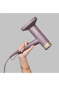 Håndholdt hårføner med en matt lavendelfinish, gullakenter, avsmalnende dyse og teksturert base. Ledningsbundet, holdt mot en grå bakgrunn.