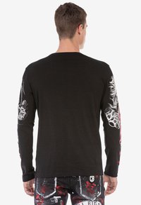 Schwarzes Langarmshirt mit weißen Grafikdesigns auf den Ärmeln. Glatte Textur mit einer insgesamt taillierten Passform.