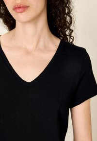 Žena s kudrnatými tmavými vlasy, která nosí černé tričko s krátkými rukávy a výstřihem do V, oříznuté tak, aby bylo vidět krk a horní část hrudníku.