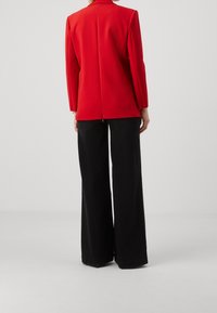 Blazer rouge ajusté avec manches longues, épaules structurées et fente au dos, associé à un pantalon noir taille haute à jambes larges.