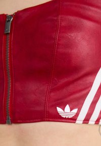 Top cropped de couro vermelho com fecho de zip, apresentando riscas brancas da Adidas e logótipo. Textura suave com forma ajustada e costuras estruturadas.