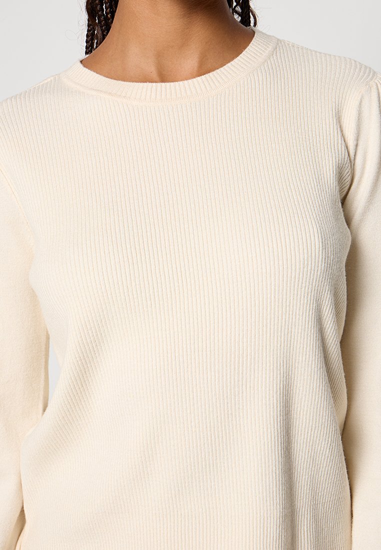 Helles beiges Rippenoberteil mit Rundhalsausschnitt, langen Ärmeln und weicher Textur, das dezente vertikale Linien über den Stoff aufweist.