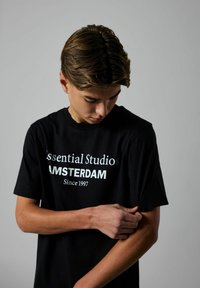 Sort bomulds T-shirt med hvid tekst, der står "Essential Studios AMSTERDAM Siden 1997." Korte ærmer, afslappet pasform.