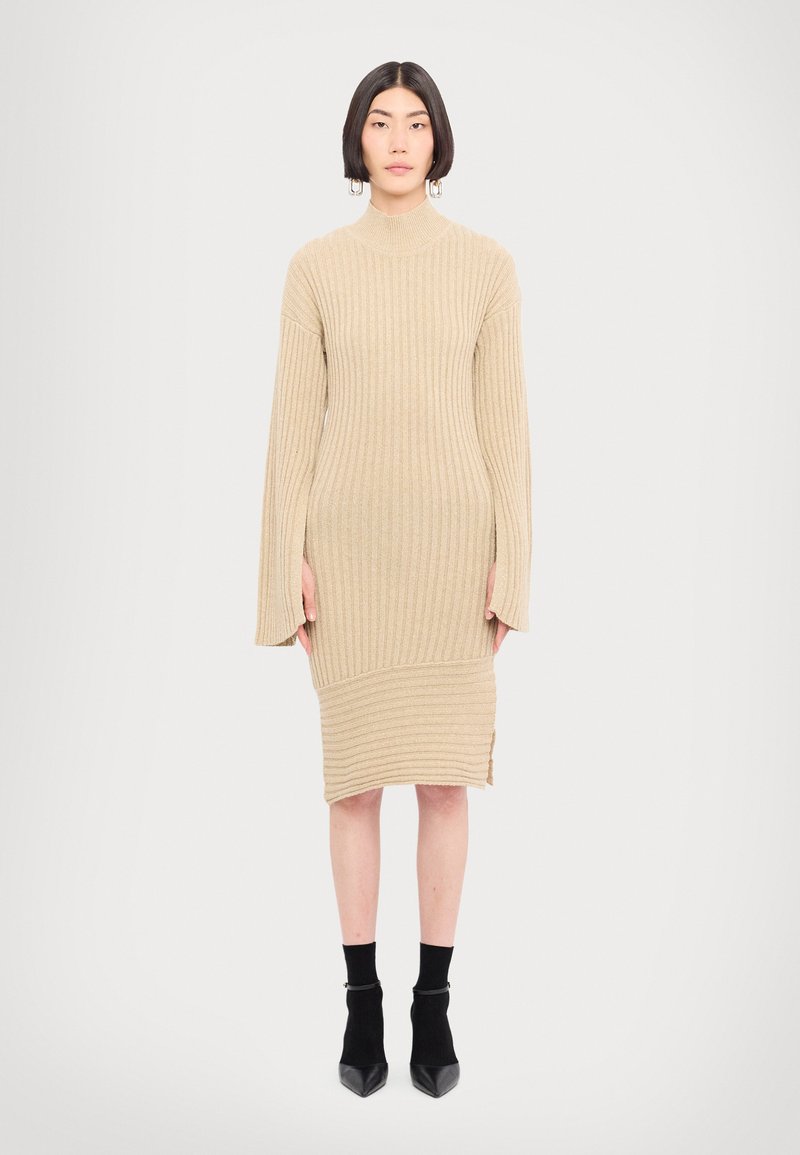 Knit beige suknelė su vientisu tekstūra, aukštu kaklu ir platėjančiomis rankovėmis. Yra šoninė plyšio detalė ir derinama su juodais aulinukais iki kulkšnies.