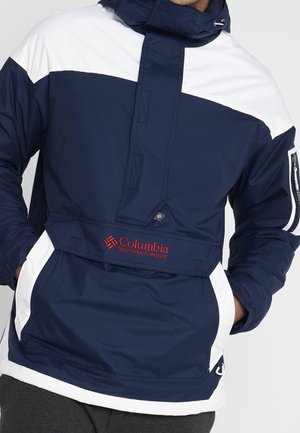Person trägt eine dunkelblaue und weiße Columbia Sportswear Kapuzenjacke mit den Händen in den vorderen Taschen.