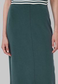 Jupe longue ajustée en vert foncé, fabriquée à partir d'un tissu doux. Elle présente une couture verticale ; des mains sont visibles tenant le tissu.