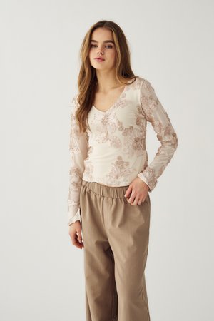 Naine, kellel on pikad pruunid juuksed, kannab pika varrukaga kreemikat pluusi lillemustriga ja lahtisi beeže pükse, seistes lihtsalt taustal.