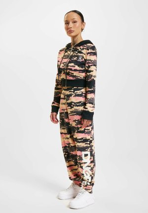 Camo-patterned jumpsuit in beige, pink, and black, featuring a zip-up hoodie, long sleeves, and a white brand logo on the leg. White sneakers.