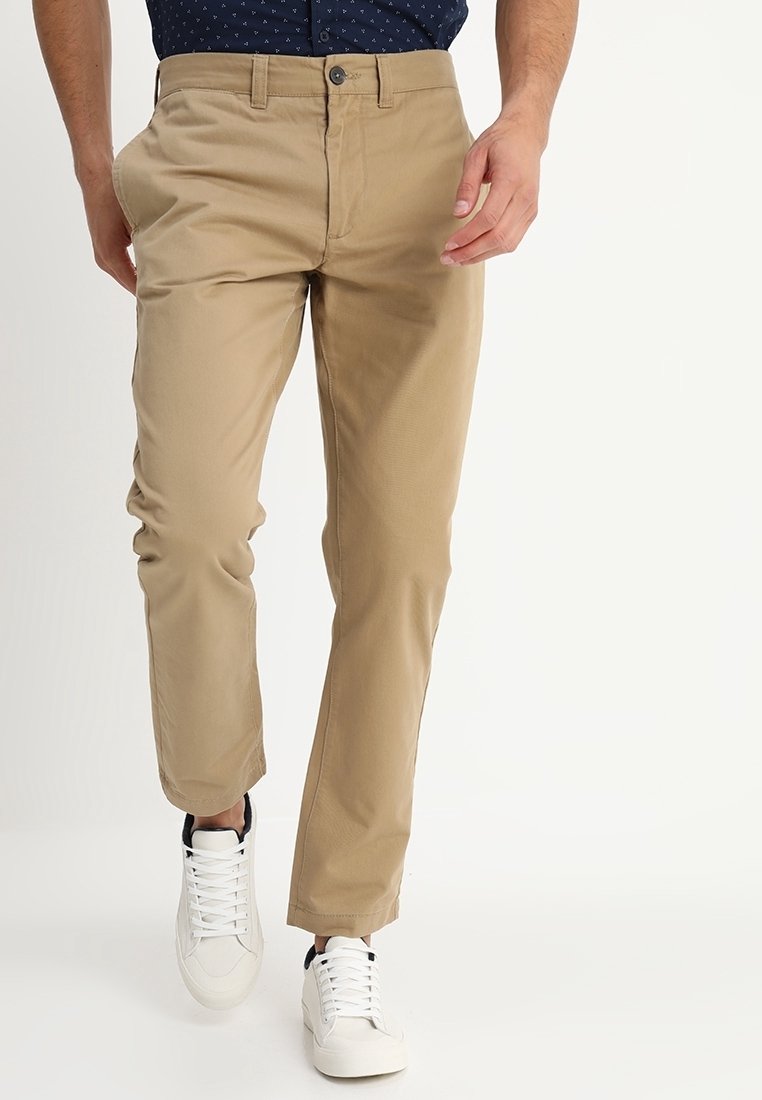 Chinos beige con taglio dritto, realizzati in tessuto misto cotone. Presentano una chiusura con bottone, passanti per la cintura e tasche laterali. Indossati con sneakers bianche.
