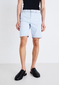 Shorts en coton bleu clair avec des revers, dotés d'une ceinture tissée blanche. Associés à des chaussures noires à enfiler en osier.