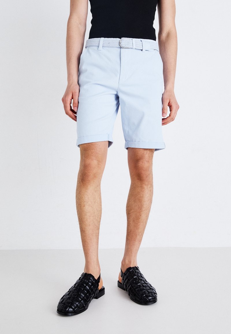 Shorts en coton bleu clair avec des revers, dotés d'une ceinture tissée blanche. Associés à des chaussures noires à enfiler en osier.