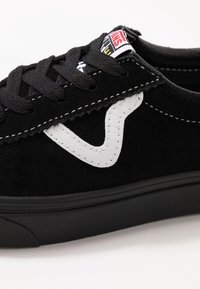 Baskets en daim noir avec lacets noirs, bande latérale blanche incurvée, semelle en caoutchouc noire et un logo Vans sur la languette.