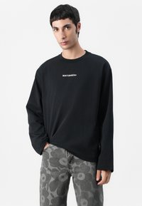 Svart langermet t-skjorte med rund hals, med hvit tekst "marimekko" sentrert på brystet. Kombinert med grå mønstrede bukser.