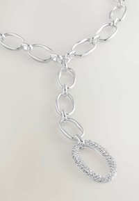 Silberkettchen mit großen ovalen Gliedern und einem Anhänger, der einen mit Strass besetzten ovalen Charm zeigt, auf einem neutralen Hintergrund.