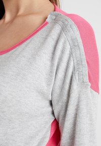 Harmaa neule, jossa on vaaleanpunaisia yksityiskohtia, leveällä kaula-aukolla ja erottuvalla hihasuunnittelulla. Pehmeä rakenne ja ribbauskohdistus olkapäällä.