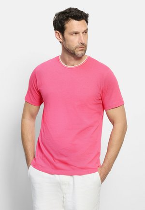 Uomo con capelli corti e scuri e barba, indossa una t-shirt rosa a maniche corte e pantaloni bianchi, in piedi con le mani in tasca, guarda verso destra.