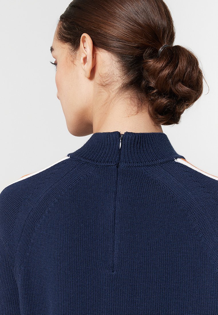 Marineblauwe gebreide trui met hoge kraag, voorzien van een rits aan de achterkant en contrasterende strepen op de schouders. Zachte textuur en aansluitend ontwerp.