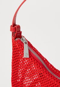 Sac en filet rouge avec une sangle en cuir rouge lisse. Dispose d'une fermeture éclair argentée avec une tirette rectangulaire. La texture est ouverte et en forme de maille.