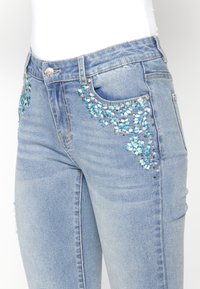 Jeans de mezclilla azul claro con un intrincado bordado floral azul y blanco en los bolsillos delanteros. Cuenta con un cierre de botón tradicional.