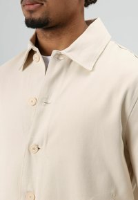 Licht beige jack met knopen, gemaakt van gestructureerde stof, met een klassieke kraag en vijf knopen aan de voorkant.