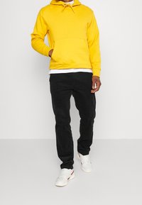 Gul huvtröja med dragsko, framficka; parat med svarta byxor och vita sneakers med röda accenter. Enkel, avslappnad design.