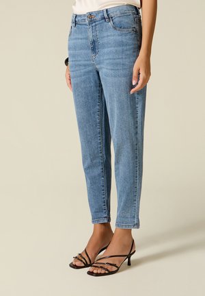 Vrouw draagt lichtblauwe high-waist slim-fit jeans en zwarte sandalen met hak en bandjes, staand op een effen achtergrond.