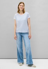 Stribet kortærmet t-shirt i lys blå og hvid, kombineret med vidde ben lys blå jeans. Modellen står i afslappede sneakers foran en grå baggrund.