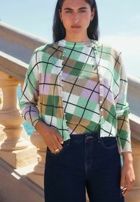 Le cardigan tricoté présente un motif argyle vert, lavande et marron, avec une fermeture à boutons et des poignets côtelés, associé à un jean noir.