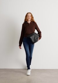 Brown, ribbed knit sweater with a zip collar, blue skinny jeans, and white sneakers, holding a black leather bag. Plain background.