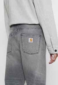 Vaqueros de mezclilla gris con un lavado sutil y costuras en contraste, con un parche del logo amarillo de Carhartt en el bolsillo trasero.