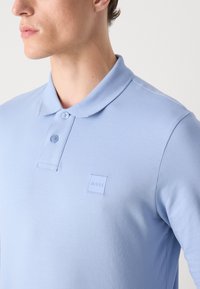 Svijetloplava polo majica s teksturiranim materijalom, dva gumba na ovratniku i kvadratna zakrpa s logom na lijevoj strani prsnog područja.
