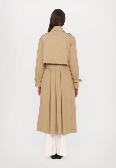 Cappotto trench color khaki con design corto, retro plissettato e cintura. Presenta maniche lunghe e colletto classico. Indossato sopra pantaloni bianchi e scarpe nere.