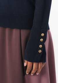 Granatowy sweter dziany z ściągaczami; złote guziki w kształcie lwa zdobią rękaw. Uzupełniony gładką spódnicą w kolorze mauve.