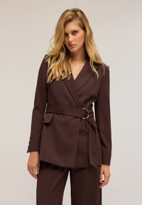 Blazer marrone su misura con scollatura a V, vita aderente e dettaglio della cintura. Realizzato in tessuto morbido, presenta tasche laterali e maniche lunghe.