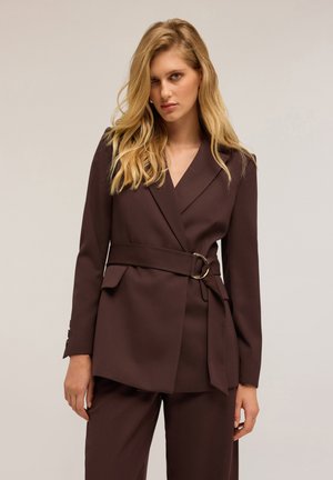 Blazer marrone su misura con scollatura a V, vita aderente e dettaglio della cintura. Realizzato in tessuto morbido, presenta tasche laterali e maniche lunghe.