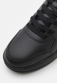 Svart läder-sneaker med perforerad tå, svarta platta snören och strukturerad gummisula. Har detaljerad sömnad och en slät yta.