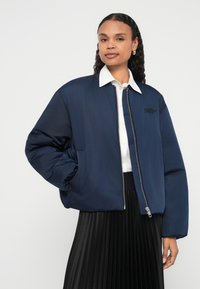 Navy puffer jacket with zip closure, side pockets, and ribbed cuffs. Features embroidered logo and paired with a white collared shirt.