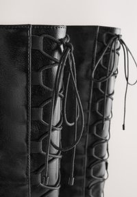 Stivali neri alti fino al ginocchio realizzati in materiale sintetico liscio con un design con lacci, caratterizzati da cuciture rinforzate e una texture sottile.