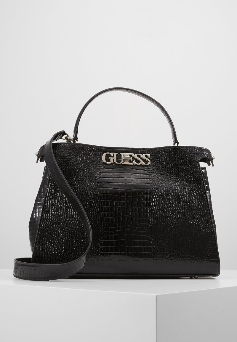 Sac à main noir embossé effet croco avec poignée supérieure, longue bandoulière et logo "GUESS" argenté à l'avant, exposé sur un socle blanc.
