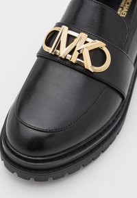 Mocassino in pelle nera con un prominente logo dorato MK su una fascia, con suola in gomma testurizzata e parte superiore liscia.