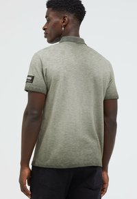 Olivgrünes Poloshirt aus strukturiertem Stoff. Kurzärmelig mit einem Markenpatch am linken Arm. Verfügt über einen klassischen Kragen.